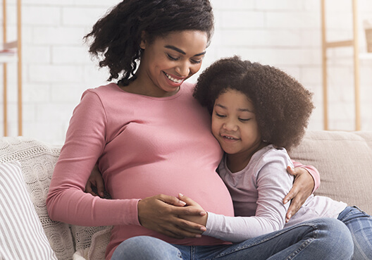 A pregnant woman wearing blue jeans and a pink top spends time with her daughter at home.