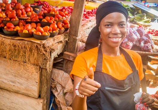 A woman running a fruit stall smiles after attending free financial education training. A woman wearing an orange t-shirt and black apron smiles and gives a thumbs up to the camera.